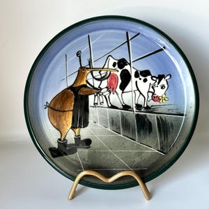 May include: A decorative ceramic plate with a whimsical design. The plate features a kiwi bird in a black coat and boots, facing two cows. The background is a blue sky with a green border. The plate is displayed on a gold-colored stand.