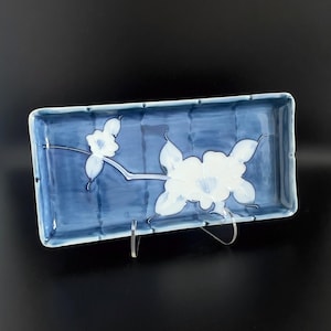 Puede incluir: Una bandeja de servicio rectangular de cerámica con un acabado esmaltado en azul profundo y un sutil patrón de cuadrícula. El centro está decorado con un motivo floral blanco pintado a mano de dos flores sobre un tallo fino, estilo pincelada.