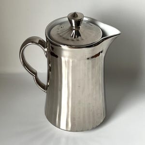 Könnte beinhalten: Eine polierte silberne Kanne mit Deckel und gebogenem Griff. Die Kanne hat einen Ausguss zum Ausgießen und einen kleinen Knopf oben auf dem Deckel. Die Oberfläche der Kanne ist reflektierend, und das Design ist schlicht und elegant.
