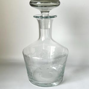May include: A clear glass decanter with a decorative stopper. The decanter has a wide base, a slender neck, and a bulbous body with etched floral designs. The stopper is a rounded shape with horizontal grey stripes.