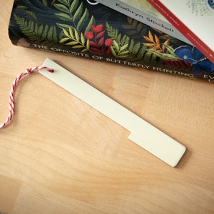 Può includere: Un segnalibro color crema con un cordino ritorto rosso e bianco. Il segnalibro è rettangolare con un'estremità dentellata. Libri sullo sfondo su una superficie di legno chiaro.