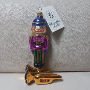 May include: A decorative Christmas ornament shaped like a toy soldier playing a golden tuba. The soldier wears a blue hat, purple jacket, and striped pants. The ornament has a tag that reads "Christopher Radko".