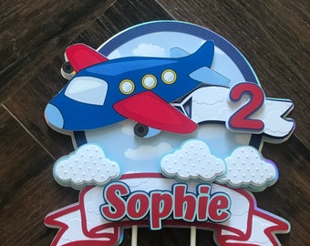 May include: Blue and red airplane cake topper with white clouds and a banner that says "Sophie" and the number "2".