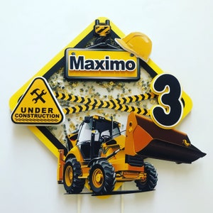 May include: A yellow and black construction themed cake topper with a yellow excavator, a sign that says "Under Construction", a sign that says "Maximo", and the number 3.