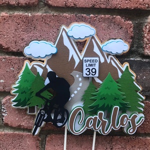 May include: A brown and white cake topper with a mountain scene, a silhouette of a person riding a mountain bike, two green pine trees, a speed limit sign that says "Speed Limit 39", and the name "Carlos" in brown letters.
