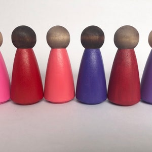 May include: Six wooden peg dolls with painted faces and different coloured dresses. The dolls are standing in a row on a white surface. The dresses are pink, red, pink, purple, red, and purple.