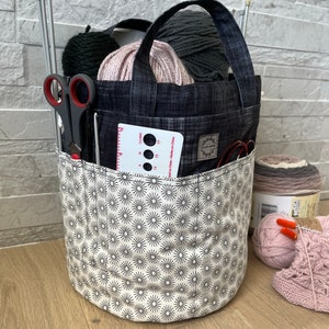 Peut inclure: Un sac de projet de tricot cylindrique avec une poignée, rempli de fil, d'aiguilles à tricoter et de ciseaux. Le sac a une poche avec un outil de jauge. Le sac est orné d'un motif d'éclat d'étoile noir et blanc sur la moitié inférieure.