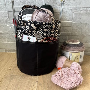 Peut inclure: Un sac en tissu à motif noir et blanc avec plusieurs poches, rempli de pelotes de laine et d'aiguilles à tricoter. Le sac est posé sur un sol en bois avec un bonnet en crochet rose et une pelote de laine devant lui.