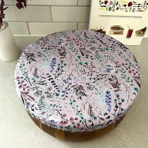 May include: A pink reusable food wrap with a floral pattern featuring various shades of purple, blue, and green. The wrap is covering a wooden bowl.