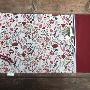 May include: A white fabric cutlery roll with a red floral pattern and a red zippered pocket. The roll is closed and has a silver fork and spoon inside.
