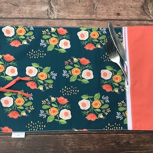 May include: A teal blue placemat with a floral pattern in shades of orange, white, and pink. The placemat has a coral coloured zippered pocket on the right side and a coral coloured tie on the left side. The placemat is made by "JoJoann's".