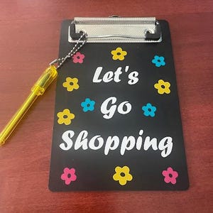 May include: Black clipboard with a white inscription "Let's Go Shopping" and colourful flower accents. A yellow pen is attached to the clipboard.
