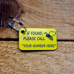 May include: A yellow rectangular keyring with a silver ring. The keyring has black text: "IF FOUND, PLEASE CALL" and "YOUR NUMBER HERE". A black bee with a heart-shaped wing is also on the keyring. The background is a wooden surface.