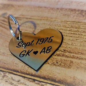 Puede incluir: Un llavero de corazón plateado con el texto "Sept 1975 GK ♥ AB" grabado en él.