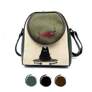 Puede incluir: Un bolso de hombro de lona verde, negro y marrón con un gato negro y un diseño de pez rojo. El bolso tiene una solapa redonda con ribete de cuero marrón.