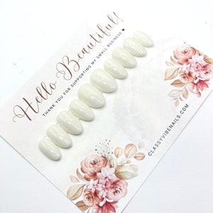 May include: A set of oval-shaped, off-white press-on nails arranged on a decorative card. The card features floral artwork in shades of pink and brown, with the text "Hello Beautiful!" and "Thank you for supporting my small business."