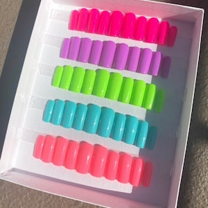 May include: A set of ten press-on nails in a white box. The nails are arranged in five rows of two, with each row featuring a different color: pink, purple, green, blue, and pink. The nails are all the same shape and size, and they have a glossy finish.