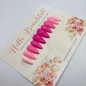 May include: A set of ten press-on nails in varying shades of pink, from light to deep fuchsia. The nails are displayed on a card with floral accents and the text "Hello Beautiful!" and "ClassyVibeNails.com".