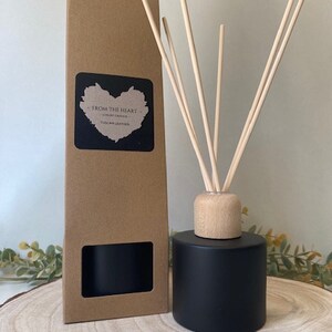 May include: A black diffuser bottle with a wooden lid and natural reeds. The bottle is sitting on a wooden surface. The bottle is labelled "From The Heart" and "Tuscan Leather".