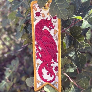 Puede incluir: Un llavero rectangular con un diseño de dragón rojo. El dragón está centrado en un fondo crema con detalles florales. Los bordes están ribeteados con hilo dorado, y un llavero dorado está sujeto en la parte superior. El llavero cuelga de una rama frondosa.