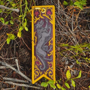 May include: A rectangular embroidered fabric keyring featuring a dark purple background with a grey wolf illustration. The design includes intricate yellow swirling vine patterns and small red floral accents, finished with a gold metal ring.