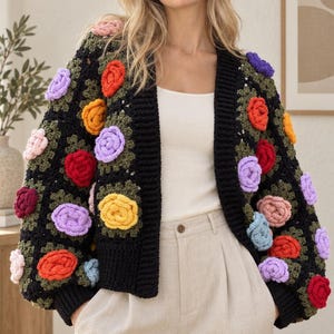 Puede incluir: Un cárdigan de ganchillo negro con un patrón de cuadrados de la abuela con centros verde oscuro y aplicaciones florales tridimensionales en tonos rojo, naranja, amarillo, azul claro, lavanda y rosa, con puños y bordes acanalados en negro.