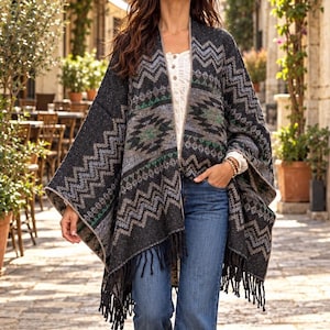May include: A black and gray open-front poncho featuring a geometric chevron pattern with green and white accents, finished with a fringed hem. The garment is worn over a white button-down shirt and blue denim flare jeans with brown suede shoes.