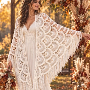 Puede incluir: Un vestido largo de color blanco roto con escote en V y detalles de encaje, combinado con un gran chal de ganchillo color crema con intrincados patrones circulares y largos flecos en los bordes, llevado sobre los hombros.