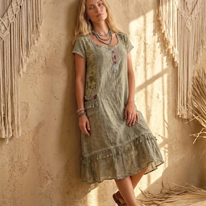 May include: A sage green knee-length dress with short sleeves, a scoop neckline, and a ruffled hem. The dress features a floral patterned side panel, a small buttoned placket at the chest, and decorative tassels along the pockets and hemline.