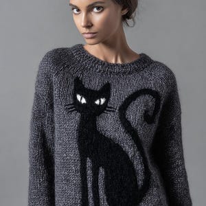 May include: A gray knit sweater featuring a black cat design. The cat is sitting with its tail curled, and has white eyes. The sweater has a crew neck and long sleeves. The background is a solid gray.