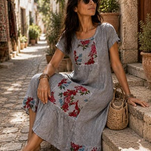 Puede incluir: Un vestido midi gris de manga corta con acabado de tela texturizada, cuello redondo y parches florales en tonos rojos y azules en el pecho, la falda y el dobladillo. El vestido tiene un corte holgado con un borde inferior con volantes y detalles.