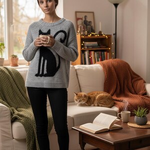 May include: A gray sweater with a black cat design. The sweater is worn with black pants and white sneakers. A woman is holding a white mug. A cat is on the couch in the background.