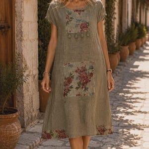 May include: A knee-length olive green dress with short sleeves and a round neckline. The garment features a rectangular floral print panel on the chest with small hanging tassels, a large floral print pocket, and a floral border at the hem.