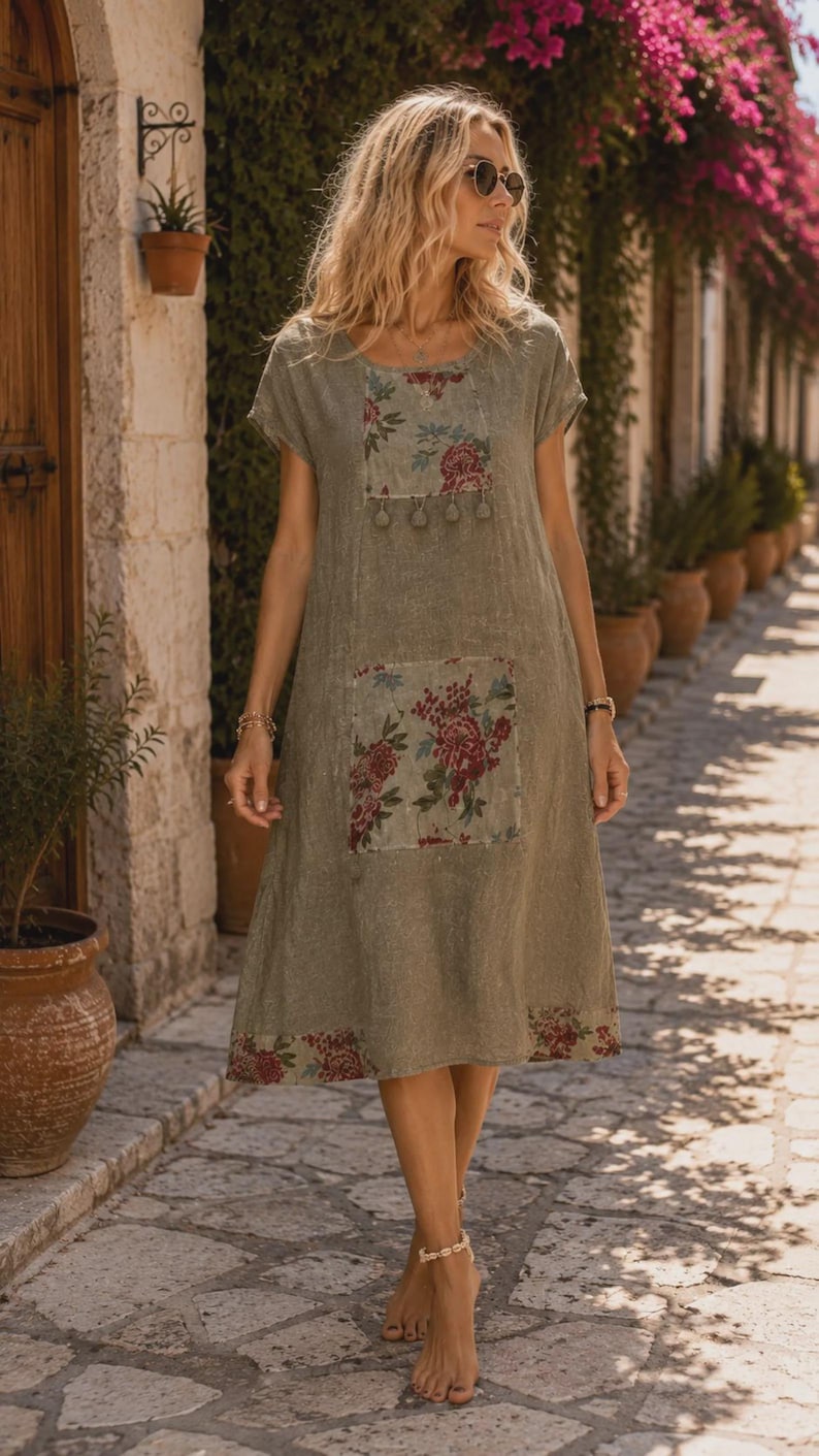 May include: A knee-length olive green dress with short sleeves and a round neckline. The garment features a rectangular floral print panel on the chest with small hanging tassels, a large floral print pocket, and a floral border at the hem.