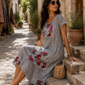 Puede incluir: Un vestido gris de largo midi con mangas cortas y cuello redondo, con parches florales en rojo, rosa y azul en el pecho, la cadera y el dobladillo. El vestido tiene un acabado de tela texturizada y un borde inferior con volantes, junto a un bolso de paja.