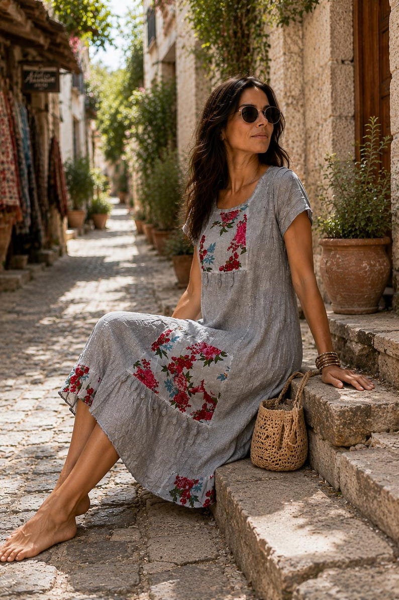 Puede incluir: Un vestido gris de largo midi con mangas cortas y cuello redondo, con parches florales en rojo, rosa y azul en el pecho, la cadera y el dobladillo. El vestido tiene un acabado de tela texturizada y un borde inferior con volantes, junto a un bolso de paja.