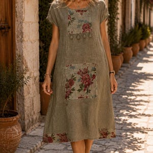 Puede incluir: Un vestido midi de color verde salvia con mangas cortas, cuello redondo y silueta holgada. La prenda presenta paneles rectangulares en el pecho y la cadera, decorados con un estampado floral en tonos rojos, blancos y verdes.