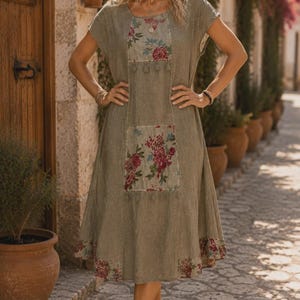 May include: A knee-length olive green dress featuring short sleeves and a round neckline. The garment is accented with rectangular panels on the chest and midsection, decorated with a floral print in shades of red, blue, and white.