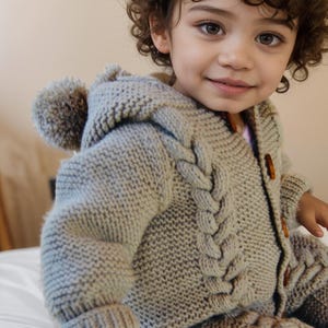 以下が含まれることがあります： ポンポンが付いたライトグレーのニットフード付きセーターを着た幼い子供。セーターはケーブルニットの模様で、ボタン留めです。