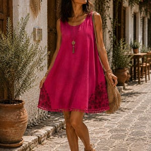 May include: A sleeveless magenta mini dress featuring a round neckline and a loose, flared silhouette. The hem is decorated with a dark floral pattern and a row of small pom-pom trim. The dress is worn with a long pendant necklace and tan sandals.