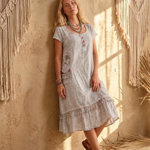 May include: A light gray knee-length dress with short sleeves, a scoop neckline, and a buttoned placket. The dress features side panels with a floral pattern, a pocket with tassel trim, and a ruffled hemline adorned with small hanging tassels.