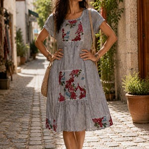 Puede incluir: Un vestido gris hasta la rodilla con mangas cortas y cuello redondo. El vestido presenta paneles rectangulares en el pecho y la falda decorados con un estampado floral en rojo, rosa y azul, rematados con peque&ntilde;as borlas grises.