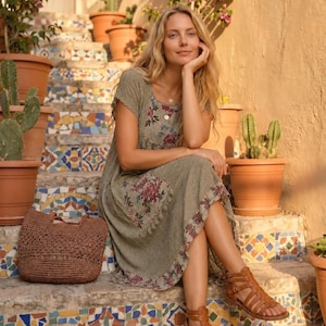 May include: A sage green midi dress featuring a floral print on the bodice, hem, and patch pocket, paired with brown strappy gladiator sandals and a woven brown tote bag. The dress has short sleeves, a round neckline, and a textured fabric finish.
