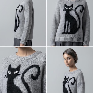 May include: A light gray sweater featuring a large black cat design on the front. The cat is depicted in a sitting pose with a curved tail. The sweater has a crew neck and long sleeves, suitable for cooler weather.