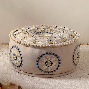 May include: A round, cream-colored ottoman with a white, yellow, and blue embroidered design. The ottoman is sitting on a white rug.