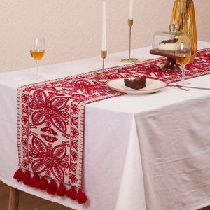 May include: A white tablecloth with a red embroidered table runner with tassels. The runner features a floral design. There are two candlesticks with white candles, two wine glasses, a plate with a piece of cake, and a glass cloche with a dessert inside.