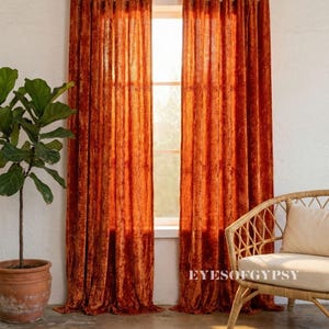 May include: Two burnt orange velvet curtains hang from a gold rod, framing a sunlit window. The floor-length curtains have a textured appearance. A potted plant and a wicker chair with a cream cushion are in the foreground.
