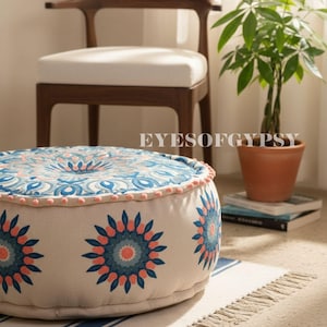 Op de afbeelding: Ronde vloerpoef met een crèmekleurige basis en een blauw en koraal bloemenpatroon. De bovenkant heeft een gedetailleerd blauw en wit ontwerp met roze rand. De poef staat op een gestreept tapijt. De tekst "EYESOFGYPSY" is zichtbaar.