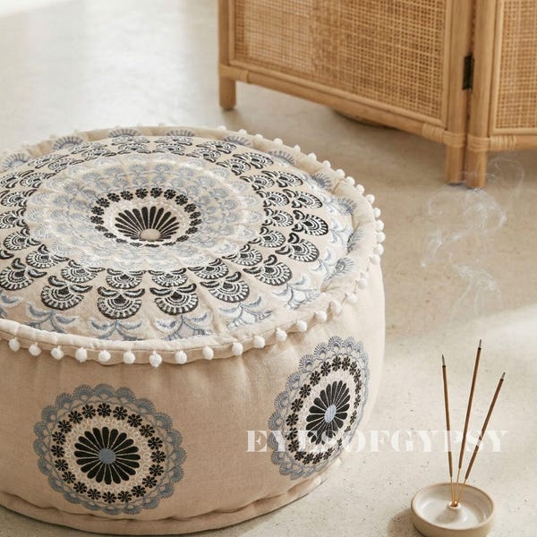 May include: A beige round floor pouf with a detailed embroidered mandala design in black and light blue. The pouf has white pom-pom trim. Incense sticks are burning nearby. The text "EYE OF GYPSY" is visible on the side of the pouf.