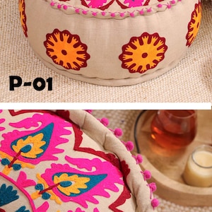 May include: A round, beige, embroidered pouf with pink, yellow, and blue floral designs. The pouf has a fringe border and is decorated with small, white pom-poms.